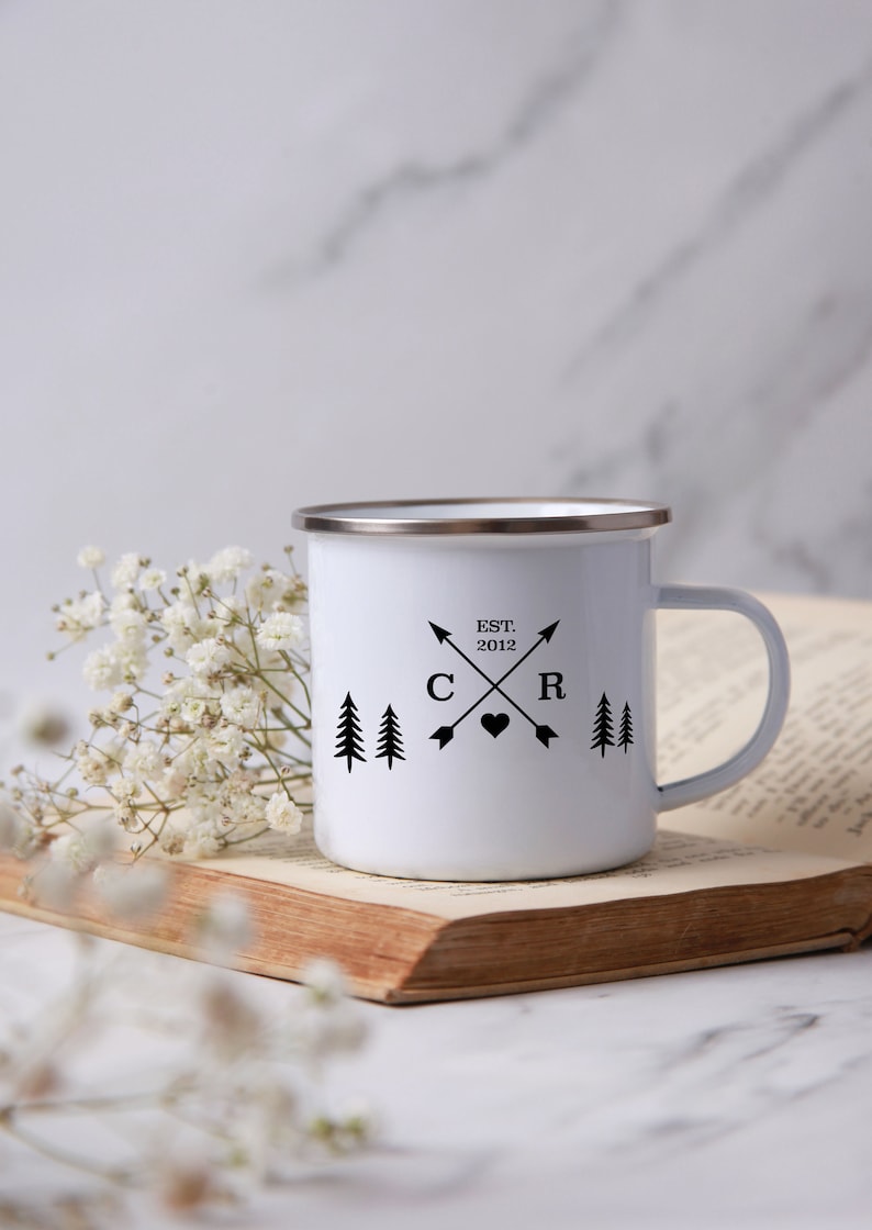 May include: White enamel camping mug with a black design featuring two pine trees, a heart, and the letters "C" and "R" with crossed arrows. The text "EST. 2012" is above the letters.