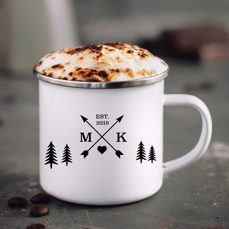 May include: White enamel camping mug with black text and graphics. The text reads "EST. 2019" with arrows pointing to the letters "M" and "K" and a heart symbol between them. There are two pine trees on each side of the design.