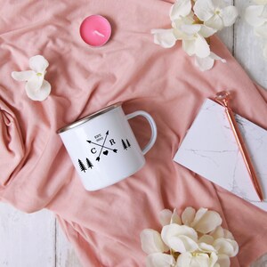 May include: White enamel camping mug with black lettering that reads "EST. 2012" and "C & R" with arrows and a heart. The mug is on a pink fabric background with white flowers.