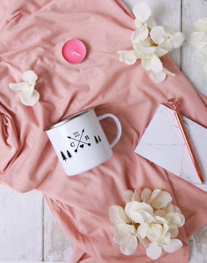 May include: White enamel camping mug with black lettering that reads "EST. 2012" and "C & R" with arrows and a heart. The mug is on a pink fabric background with white flowers.