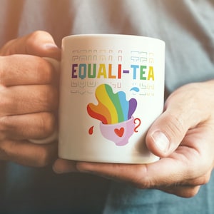 Pode incluir: Caneca de cerâmica branca com um design de arco-íris e o texto "EQUALI-TEA". O arco-íris está derramando de uma xícara de chá rosa com um coração vermelho.