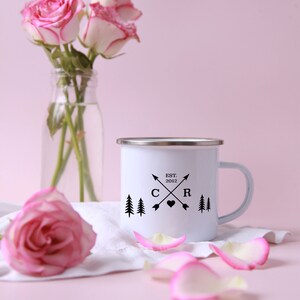 May include: White enamel mug with a black design featuring two pine trees, a heart, and the text "EST. 2012" with the letters "C" and "R" on either side of the heart. The mug has a silver rim and handle. The mug is sitting on a white cloth with pink rose petals.