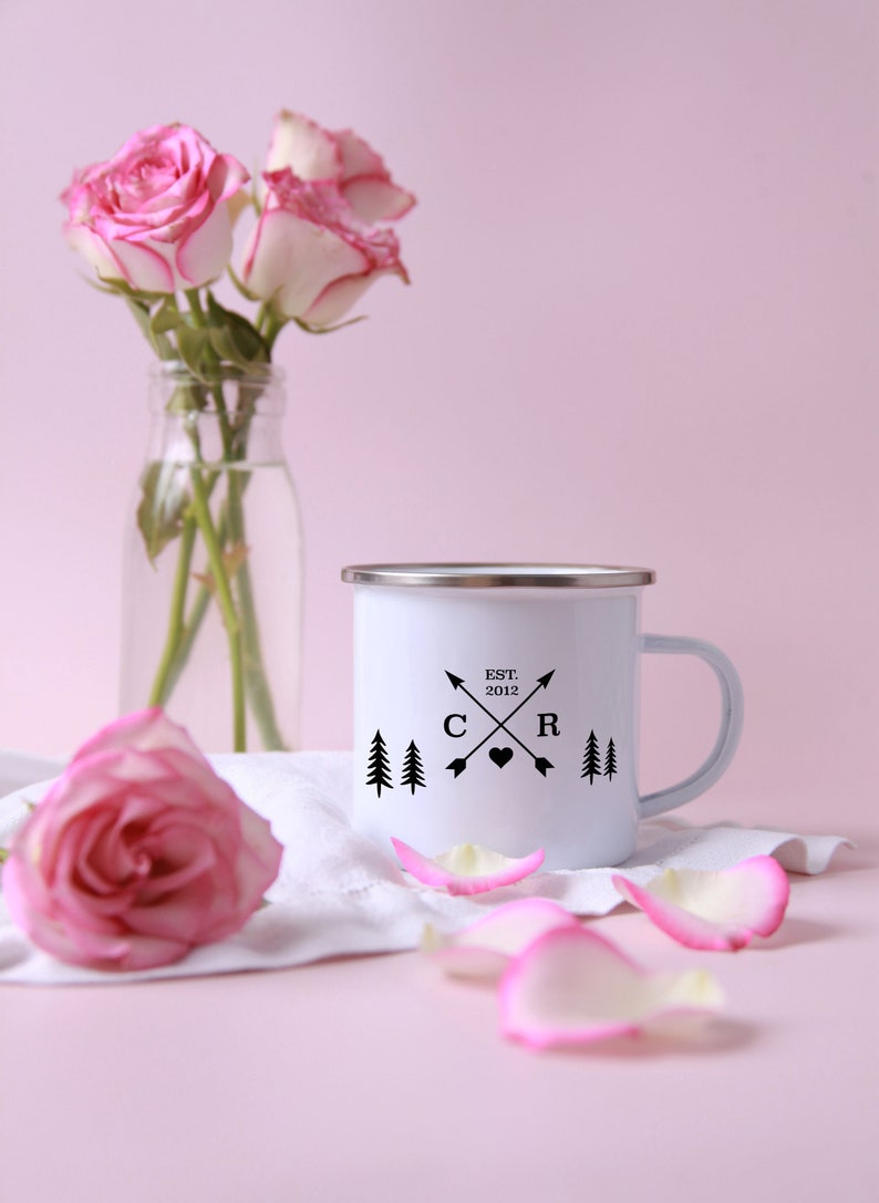 May include: White enamel mug with a black design featuring two pine trees, a heart, and the text "EST. 2012" with the letters "C" and "R" on either side of the heart. The mug has a silver rim and handle. The mug is sitting on a white cloth with pink rose petals.