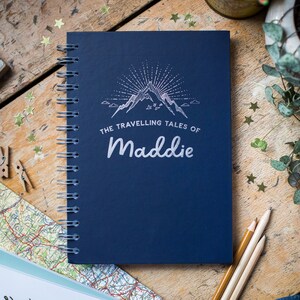 Può includere: Un taccuino blu navy rilegato a spirale con la scritta "The Travelling Tales of Maddie" in bianco. La copertina presenta un disegno di catena montuosa con linee radianti. Il taccuino è su una superficie di legno con matite e coriandoli a forma di stella.