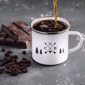 May include: White enamel camping mug with black lettering that reads "EST. 2011 C R" with a heart and arrows between the letters. Two pine trees are on either side of the letters. Coffee is being poured into the mug.