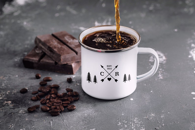 May include: White enamel camping mug with black lettering that reads "EST. 2011 C R" with a heart and arrows between the letters. Two pine trees are on either side of the letters. Coffee is being poured into the mug.
