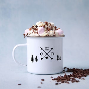 May include: White enamel mug with a black design of two trees, arrows, and the letters "C" and "R" with a heart in between. The text "EST. 2012" is above the letters. The mug is filled with hot chocolate topped with whipped cream, marshmallows, and chocolate sprinkles.