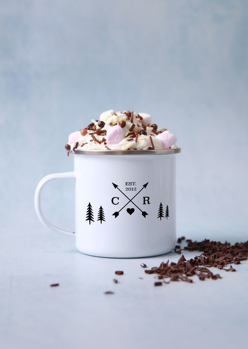 May include: White enamel mug with a black design of two trees, arrows, and the letters "C" and "R" with a heart in between. The text "EST. 2012" is above the letters. The mug is filled with hot chocolate topped with whipped cream, marshmallows, and chocolate sprinkles.