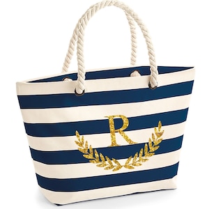 May include: A white and navy blue striped tote bag with a gold glitter initial "R" and a gold glitter laurel wreath. The bag has rope handles.