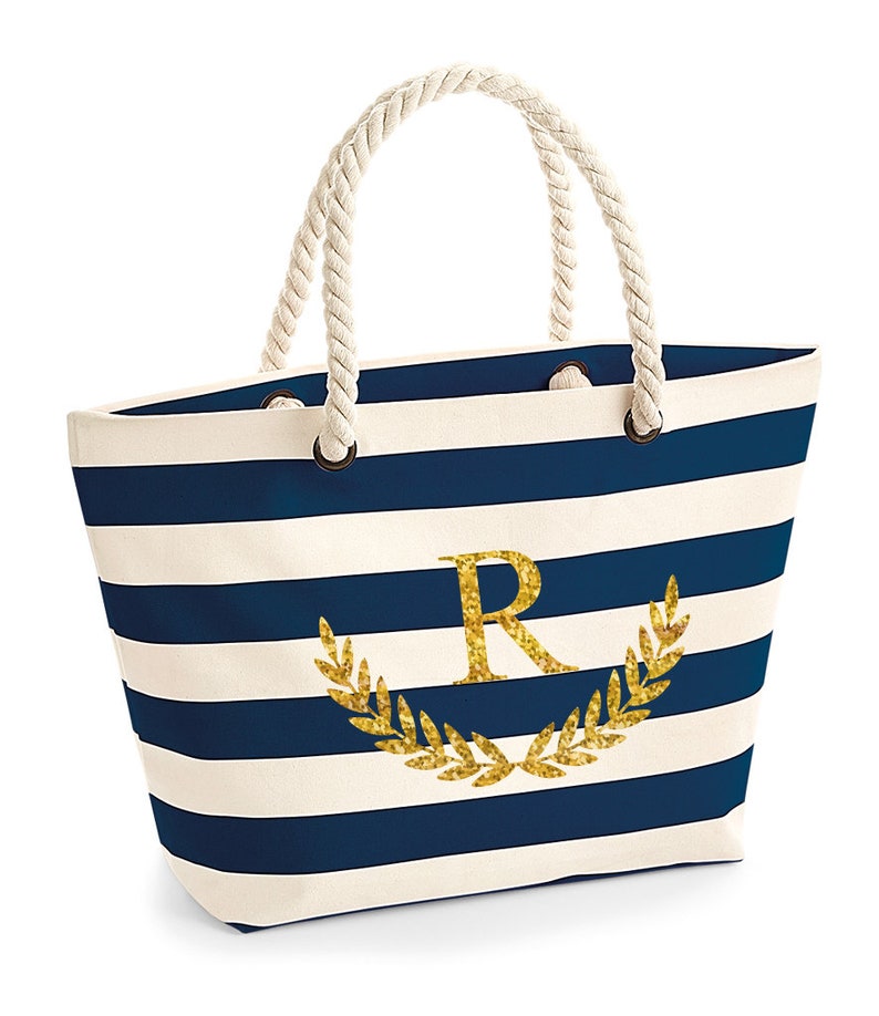 May include: A white and navy blue striped tote bag with a gold glitter initial "R" and a gold glitter laurel wreath. The bag has rope handles.