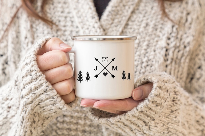 May include: White enamel mug with black lettering and a design of two crossed arrows with a heart in the center. The arrows have the letters J and M at the ends. The text "EST. 2019" is above the arrows. There are three small pine trees on each side of the design.