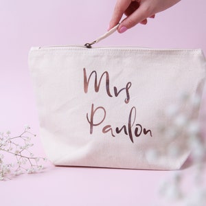 May include: A white canvas makeup bag with a zipper closure. The bag has the words "Mrs. Panlon" printed in rose gold foil.