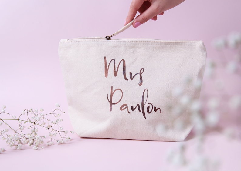 May include: A white canvas makeup bag with a zipper closure. The bag has the words "Mrs. Panlon" printed in rose gold foil.