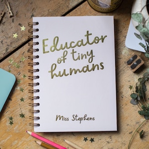 Op de afbeelding: Een wit spiraalgebonden notitieboek met gouden letters met de tekst "Educator of tiny humans" en "Miss Stephens". Het notitieboek ligt op een houten oppervlak, met potloden en stervormige confetti.