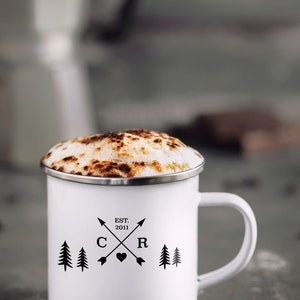 May include: White enamel mug with a black design of two pine trees, a heart, and the letters "C" and "R" with the text "EST. 2011" above. The mug is filled with a cappuccino with a brown foam top.