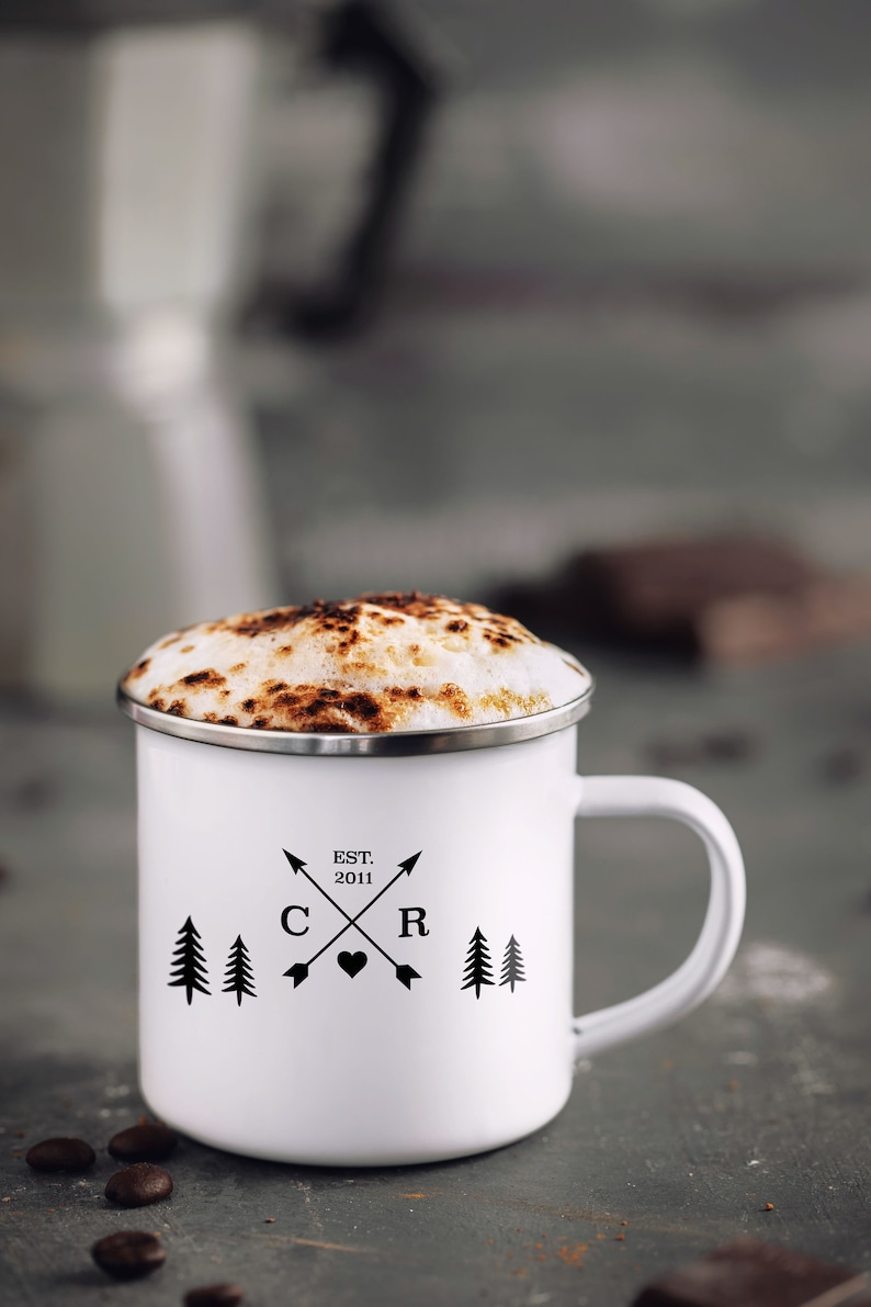 May include: White enamel mug with a black design of two pine trees, a heart, and the letters "C" and "R" with the text "EST. 2011" above. The mug is filled with a cappuccino with a brown foam top.