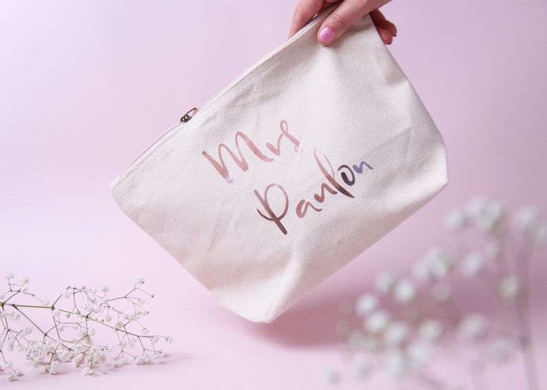 May include: A white canvas zippered pouch with the text "Mrs. Paulson" in rose gold foil lettering.