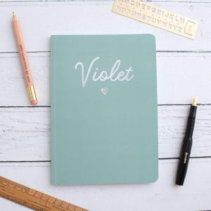Puede incluir: Un cuaderno de color verde azulado claro con el nombre "Violet" en escritura plateada y un pequeño corazón. Un lápiz rosa, una pluma estilográfica negra, una plantilla de letras dorada y una regla de madera están dispuestas alrededor del cuaderno sobre una superficie de madera blanca.