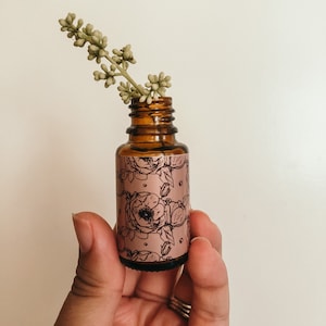 May include: A small amber glass bottle with a floral design label. The bottle is filled with a clear liquid and has a dropper top. A sprig of green foliage is placed on top of the bottle.