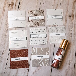 May include: Nine square labels with different patterns and text. The labels have words like "sleep", "wellness", "breathe", "brave", "calm", "relief", "headache", "energy", and "focus". A brown glass roller bottle with a gold cap and a white floral design is in the center of the image.