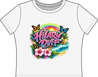Camiseta estampada de Hilary Duff con diseño de playa tropical y mariposas al atardecer.