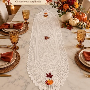May include: An oval white lace table runner featuring intricate floral patterns and small colorful autumn-themed appliqués including pumpkins and leaves. The runner includes text at the top that reads Customizable Choose your appliqués.
