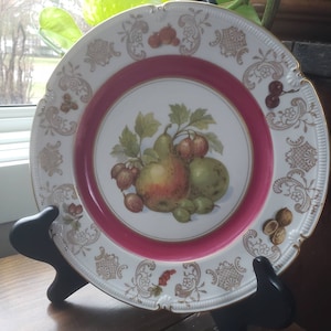 May include: A white porcelain plate with a pink rim and gold trim. The center of the plate features a painted image of pears, gooseberries, and leaves.