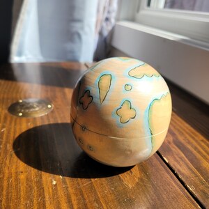 May include: A wooden sphere, hand-painted with a map-like design in shades of tan, blue, and green. The decorative globe sits on a dark wooden surface, with a thin wire encircling its middle. The sphere is approximately 4 inches in diameter.