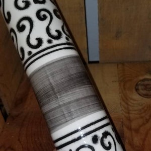 May include: A white ceramic rolling pin with black and gray decorative bands. The bands feature a repeating pattern of swirls.