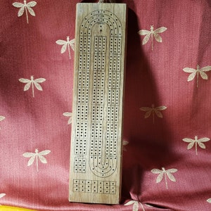 Può includere: Un tabellone da cribbage in legno con un design tradizionale. Il tabellone ha una pista per i pioli e un'area di punteggio.