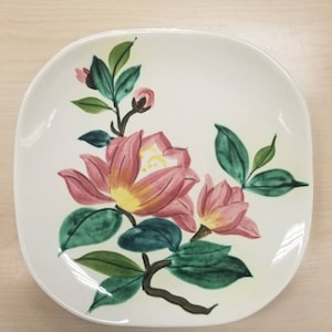 May include: White square plate with a floral design. The plate features pink and yellow flowers with green leaves and dark brown stems. The flowers are painted in a detailed, artistic style.