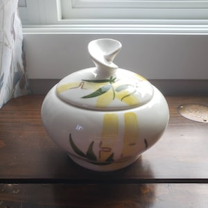 以下が含まれることがあります： 黄色と緑の花柄の白いセラミック製の砂糖入れ。蓋はユニークな渦巻きデザインです。