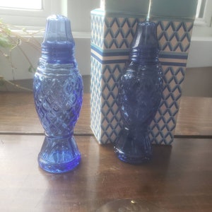 May include: Two blue glass salt and pepper shakers in the shape of fish. The shakers have a diamond pattern and are sitting on a wooden surface.