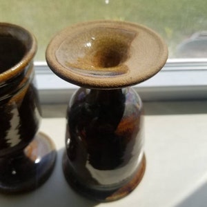 May include: Two brown ceramic mugs with a glazed finish. The mugs have a wide base and a narrow opening.