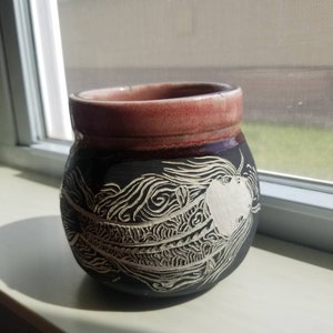 May include: A black ceramic pot with a red rim and a white design of a fish with intricate details.