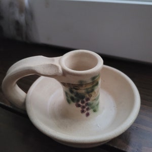 May include: A white ceramic candle holder with a handle and a grapevine design. The holder has a small, round base and a larger, round top with a hole in the center for a candle.
