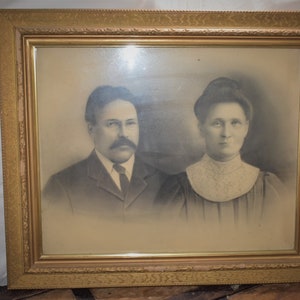 May include: A black and white portrait of a couple in a gold frame. The man is wearing a suit and tie, and the woman is wearing a dress with a lace collar.