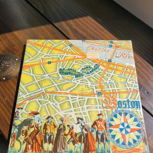 May include: A vintage game board depicting a map of Boston, Massachusetts. The board features a compass, a yellow background, and red and orange roads. The text "Charles River" and "Boston" are visible on the board.