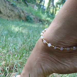 May include: Ankle bracelet featuring rose quartz beads and gold-colored accents. The delicate jewelry is displayed on an ankle, set against a backdrop of green grass. The bracelet's design is simple and elegant.