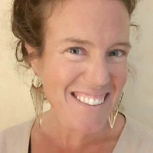 Puede incluir: Una mujer con el pelo castaño rizado sonríe y lleva un jersey beige claro. Lleva unos pendientes grandes de oro con forma geométrica.