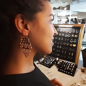 May include: A woman wearing a black top and large geometric gold earrings. She is looking at a display of jewelry, including earrings, rings, and bracelets.