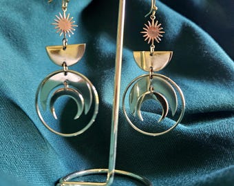 May include: A pair of celestial-themed earrings. Each earring features a sunburst, a half-moon shape, and a crescent moon within a circular frame. The earrings are displayed on a silver metal stand against a teal fabric backdrop.
