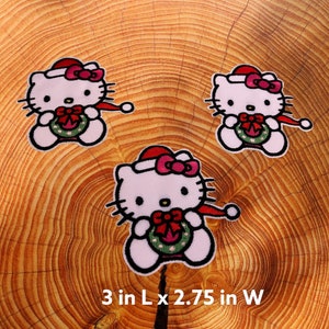 May include: Three embroidered iron-on patches featuring a cartoon cat wearing a Santa hat and holding a Christmas wreath. The patches are 7.6 cm long by 7 cm wide.