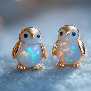 May include: A pair of gold-colored penguin earrings. The penguins have a gold head and wings, with a white body that has colorful iridescent flecks. The eyes are small and blue. The earrings are on a light blue background.