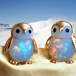 Könnte beinhalten: Zwei goldfarbene Pinguin-Ohrringe mit opalisierenden blauen und mehrfarbigen Körpern. Die Ohrringe haben schwarze Augen und sind vor einem schneebedeckten Hintergrund platziert.
