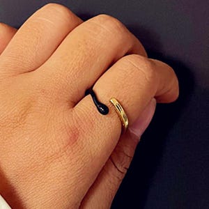 May include: A gold-colored ring with a black enamel design. The ring has an open design with a curved black enamel accent that wraps around the finger. The ring is on a person's finger.