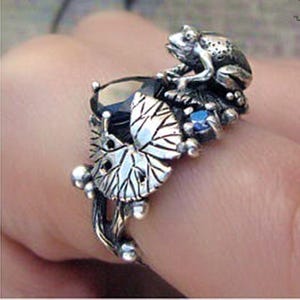 May include: A silver ring with a frog design, set with a large black gemstone and small blue stones. The ring features detailed leaf and floral accents. The frog is the main focus of the ring, with intricate detailing.