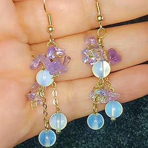 May include: Gold-tone dangle earrings featuring clusters of small, light purple gemstones and three opalescent, round beads. The earrings have a delicate chain and hook closure.