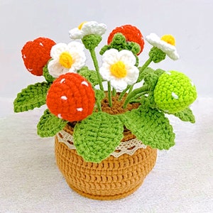 Op de afbeelding: Een gehaakte aardbeienplant met rode en groene aardbeien, witte bloemen en groene bladeren in een bruine gehaakte pot.