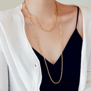 May include: A close-up shot of two gold necklaces of different lengths. The necklaces are worn over a black top and a white cardigan. The longer necklace hangs down, while the shorter one rests near the collarbone. The jewelry is simple and elegant.
